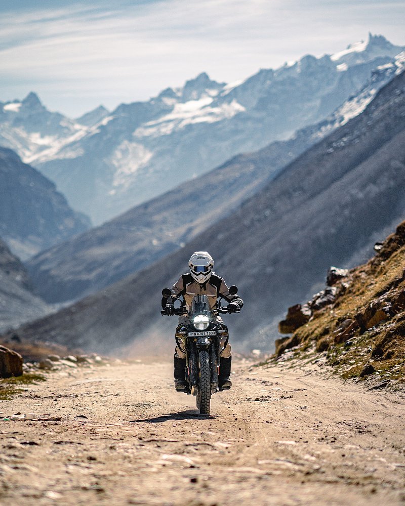 Dustin riding the Royal Enfield Himalayan on a remote dirt road along a river with the snow-covered mountains in the distance