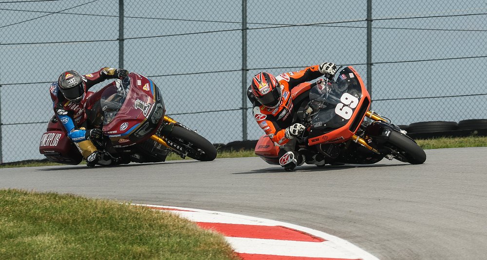 a Harley-Davidson and an Indian rider both dragging knee in a corner during a King of the Baggers race