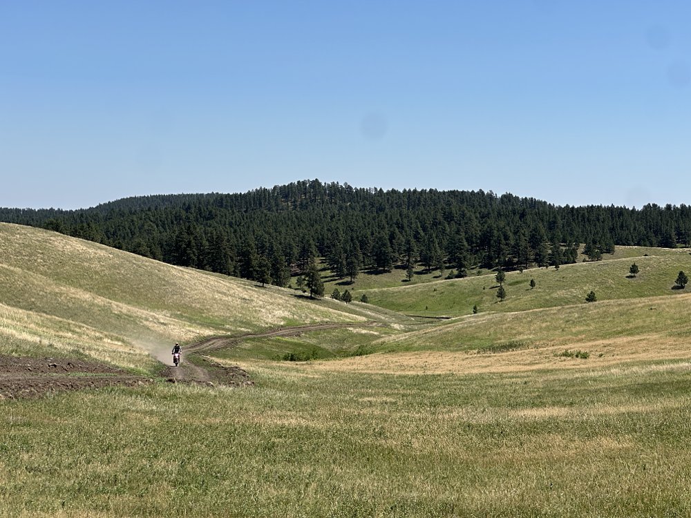 A rural dirt road running through a shallow valley with green pastures on either side, with a 2026 KTM 690 Enduro R barely visible. 