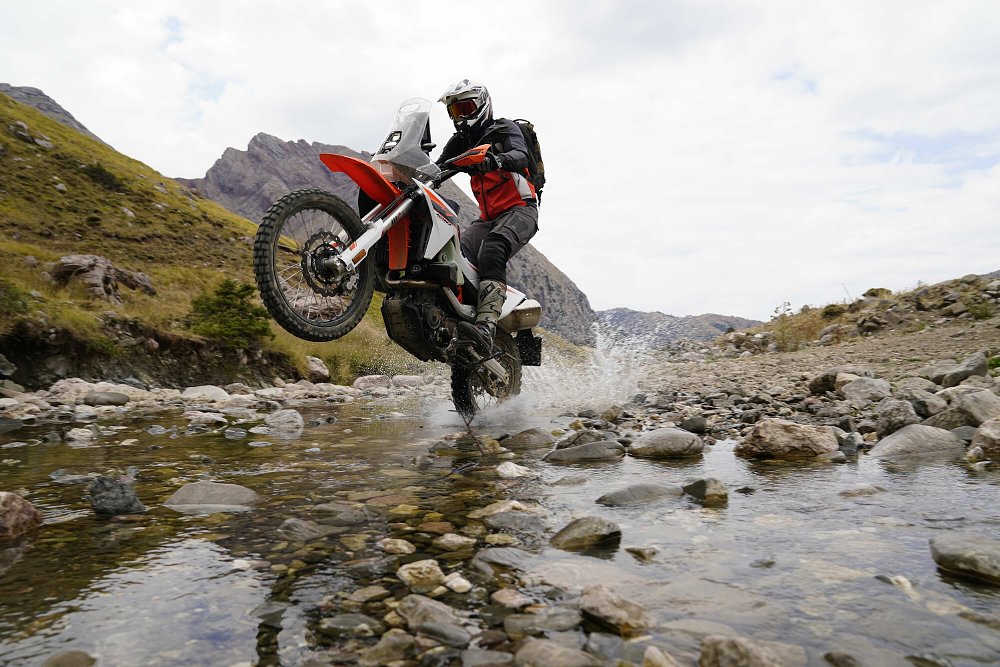 2027 KTM 690 Rally wheelying across a creek with rocky hills in the background.