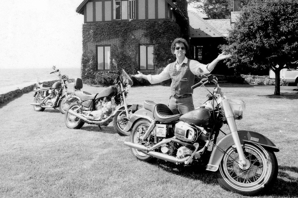 An archival image of Billy Joel posing next to three motorcycles.