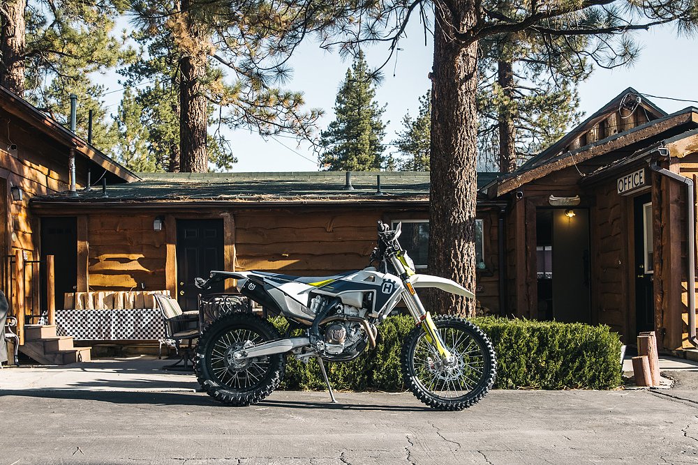 A Husqvarna motorcycle parked in front of a wood cabin and pine trees.