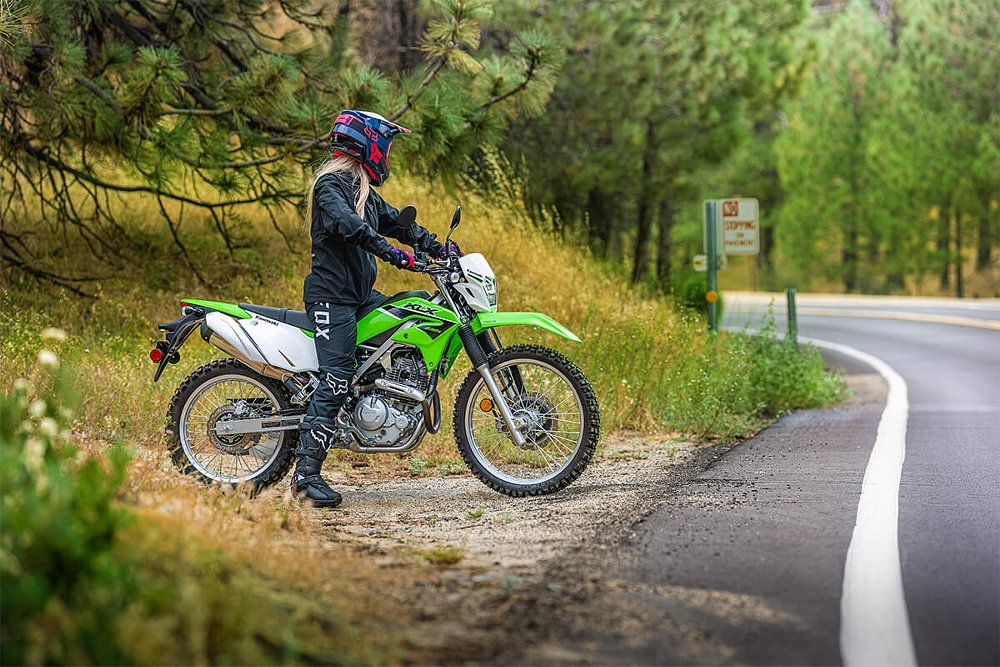 A short woman rider sits at a stop sign on a mountain road on a green kawasaki KLX230 dual sport motorcycle, with her feet flat on the groun