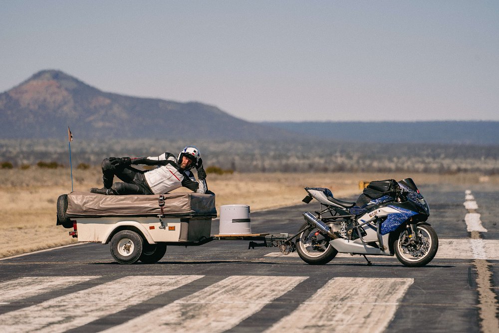 A 2006 Suzuki GSX-R1000 parked, with a camping trailer hitched to the back and a person lying on top of the trailer.