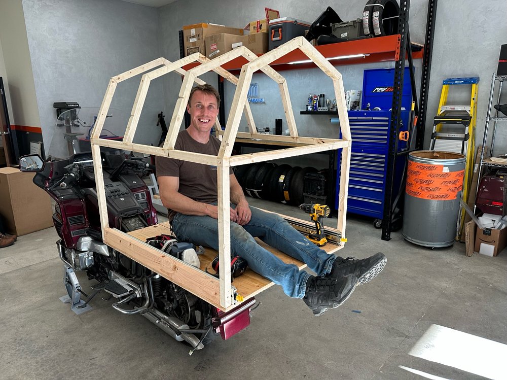 A 1989 Honda Gold Wing with the framing for a small building attached to the back, and a man sitting inside.