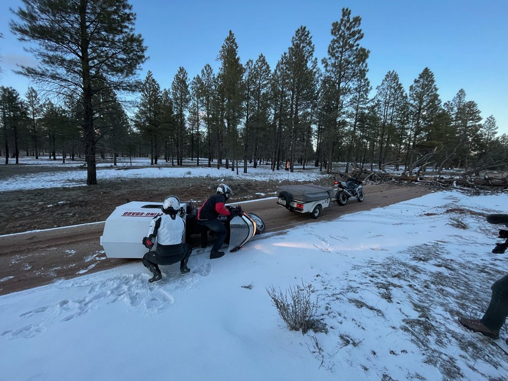 Two men picking up a motorcycle on a dirt road, with another motorcycle and a tree across the road in the background.