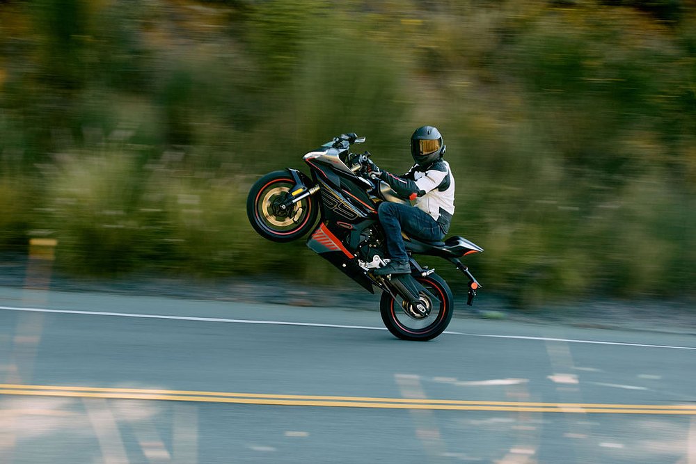 A CFMOTO 450SS sport bike does a wheelie on a rural road while the rider looks at the camera.