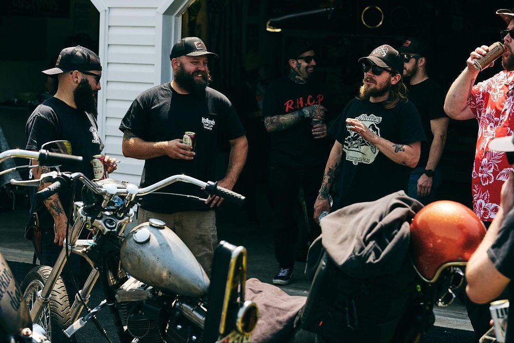 A group of bikers hanging around the garage with their bikes parked, laughing and drinking beer