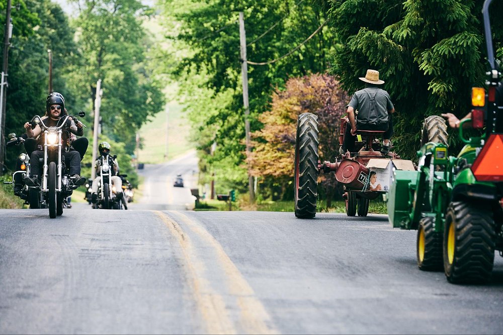 A photo of cruiser motorcycles going down a forested rural road with tractors on the otherside