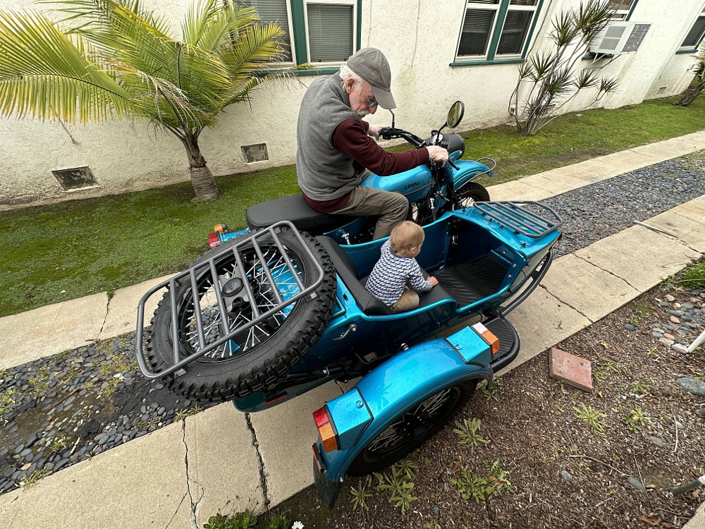 A grandfather and grandson sit in a Ural in a dirveway.