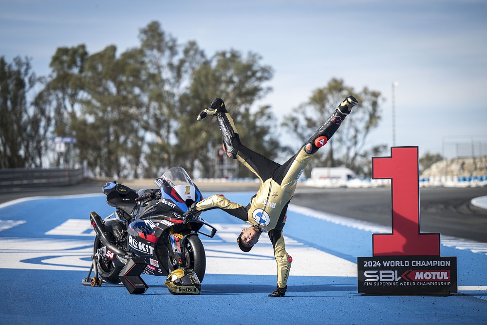 Toprak Razgatlıoğlu doing a handstand beside his motorcycle and a huge number one symbol to celebrate his 2024 championship