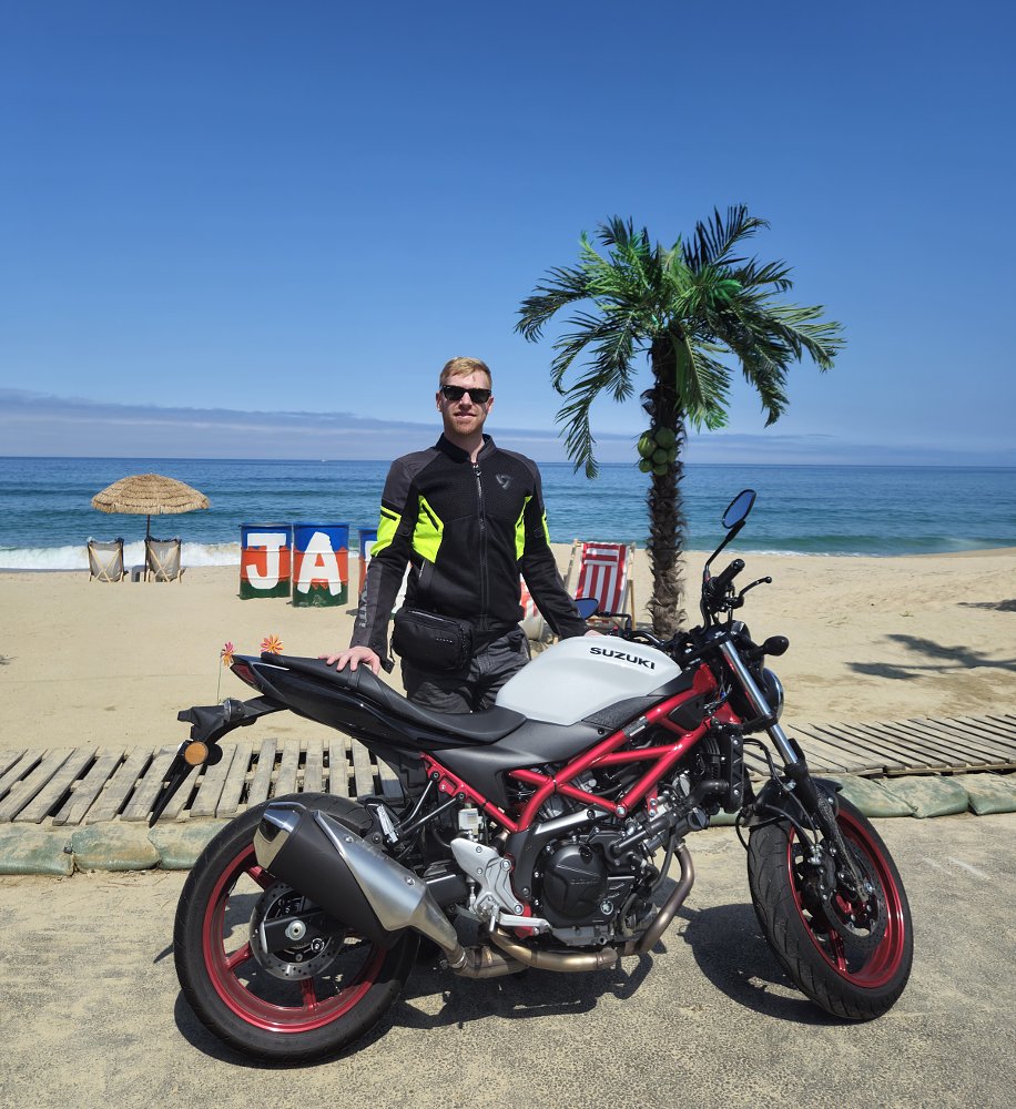 author standing behind his white Suzuki SV650 with red trellis frame and red wheels at the beach