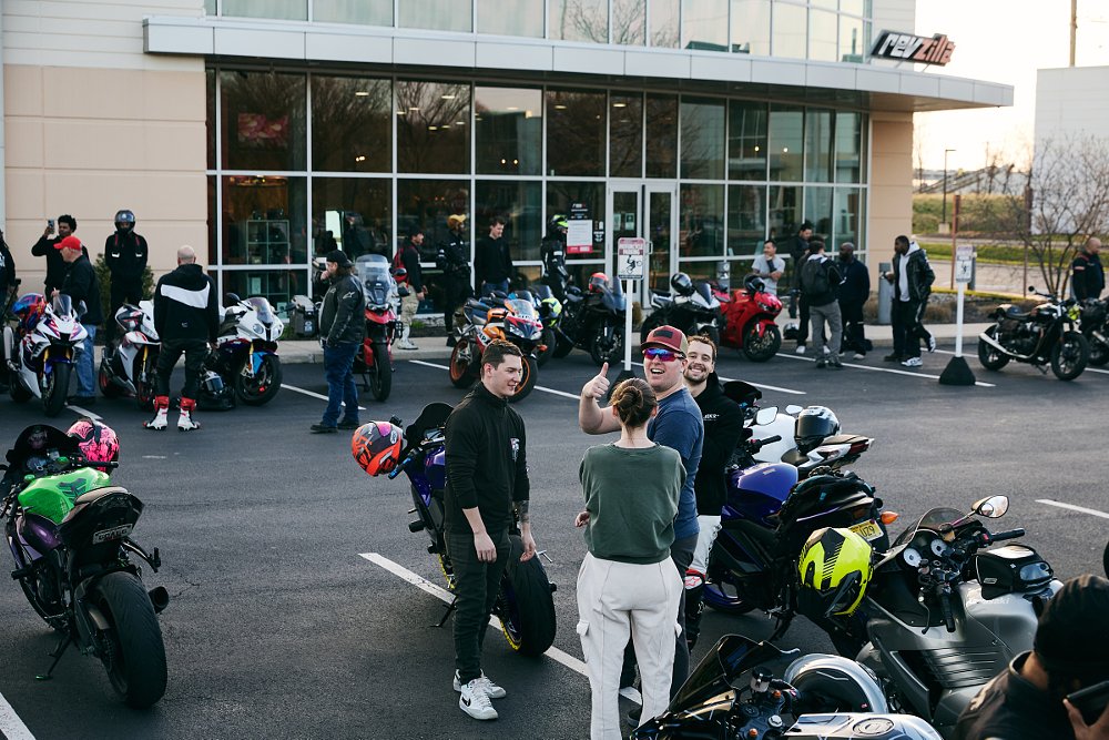 group of riders and motorcycles in the evening outside the RevZilla store, one rider smiling and waving to the camera