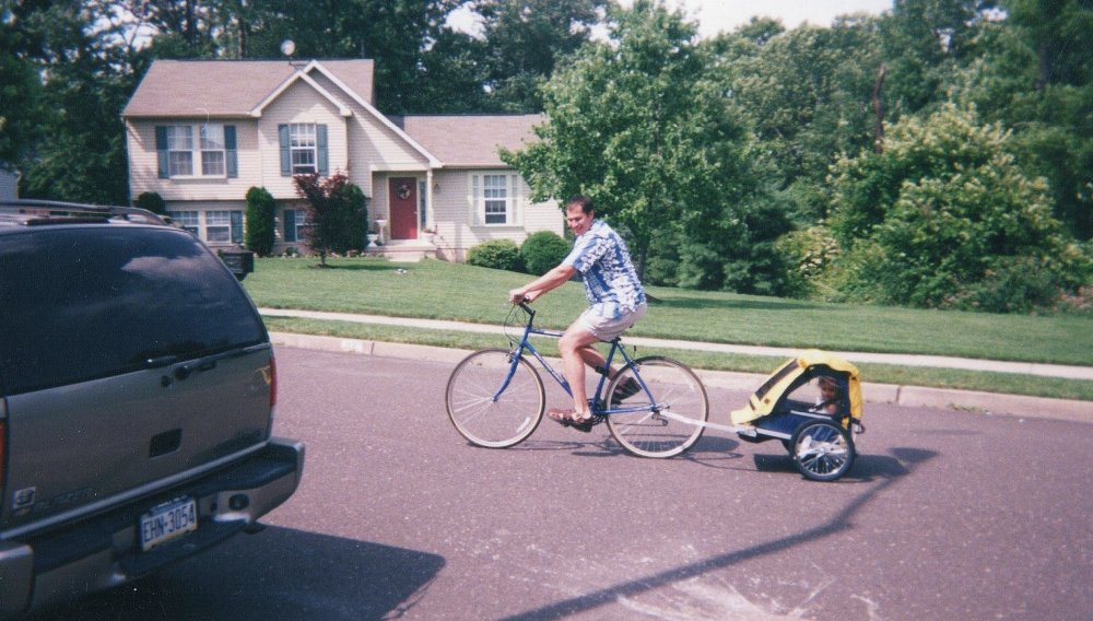 author on his bicycle on a suburban street towing his small child in a trailer behind
